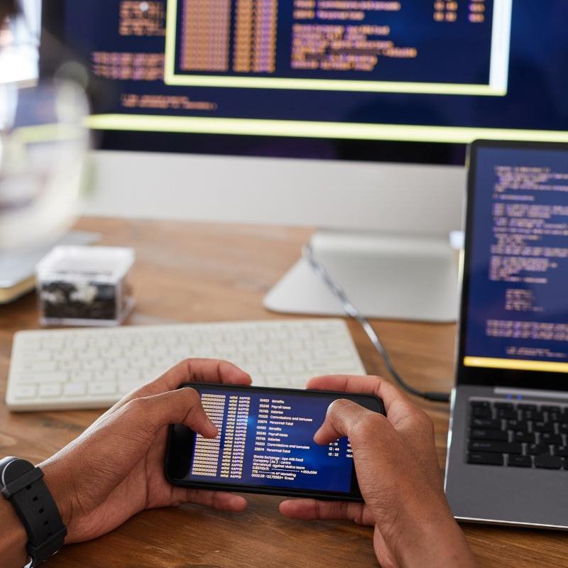 App developer holding a cell phone in front of a computer, coding for mobile app development.